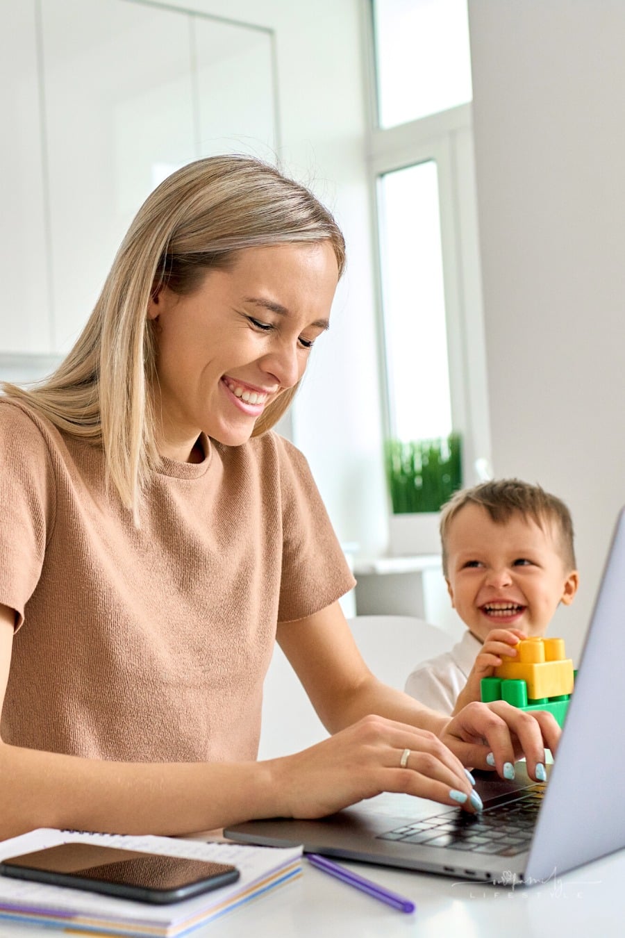 mom working at laptop while laughing with young son nearby