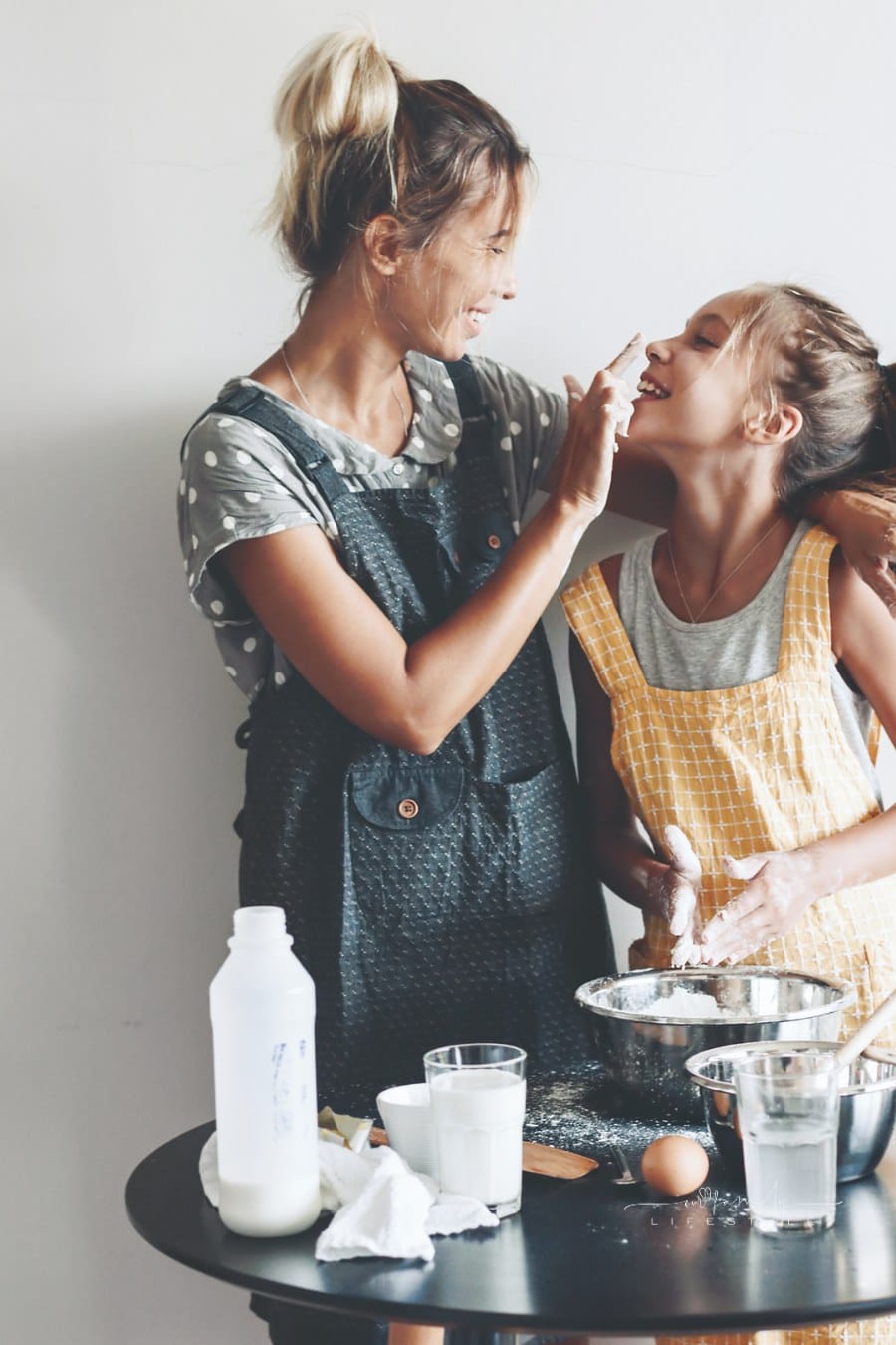 Mom with her daughter dressed in linen aprons are cooking together over light wall