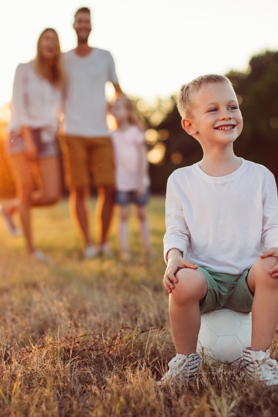 Mother, father, and daughter laughing and posing behind son sits on white soccer ball in foreground while enjoying beautiful summer day outdoors