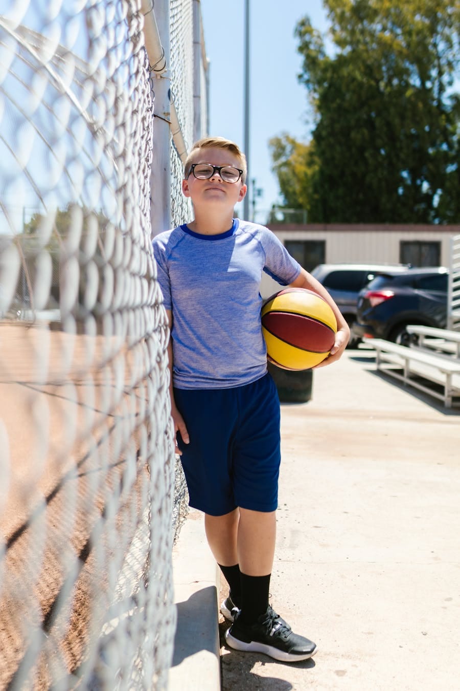 A boy leaning against a fence holding a basketball on a sunny day outdoors.