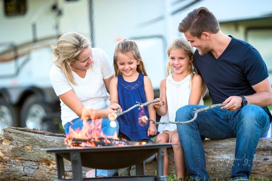 A family of four are roasting marshmallows together around a campfire while on vacation. Their RV is parked in background.