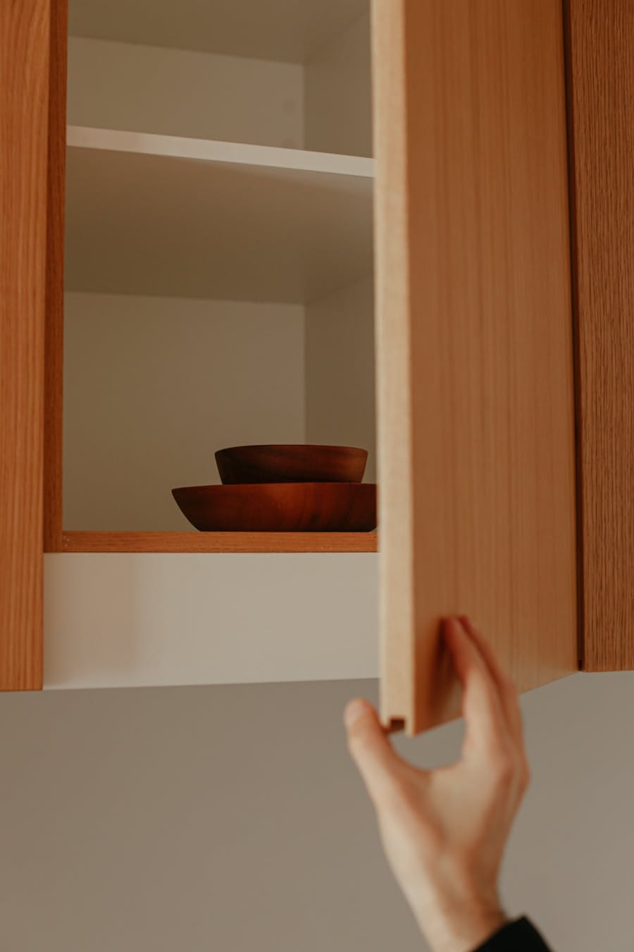 A hand opens a wooden kitchen cabinet to reveal stacked brown bowls inside.