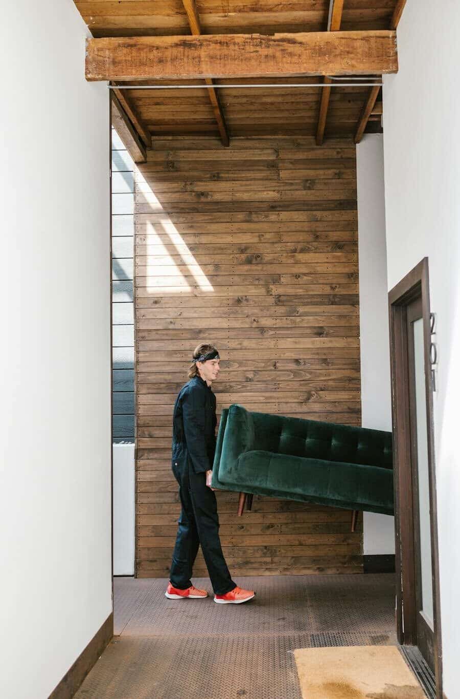 A mover carries a green sofa through a modern wooden hallway, showcasing relocation and moving service.