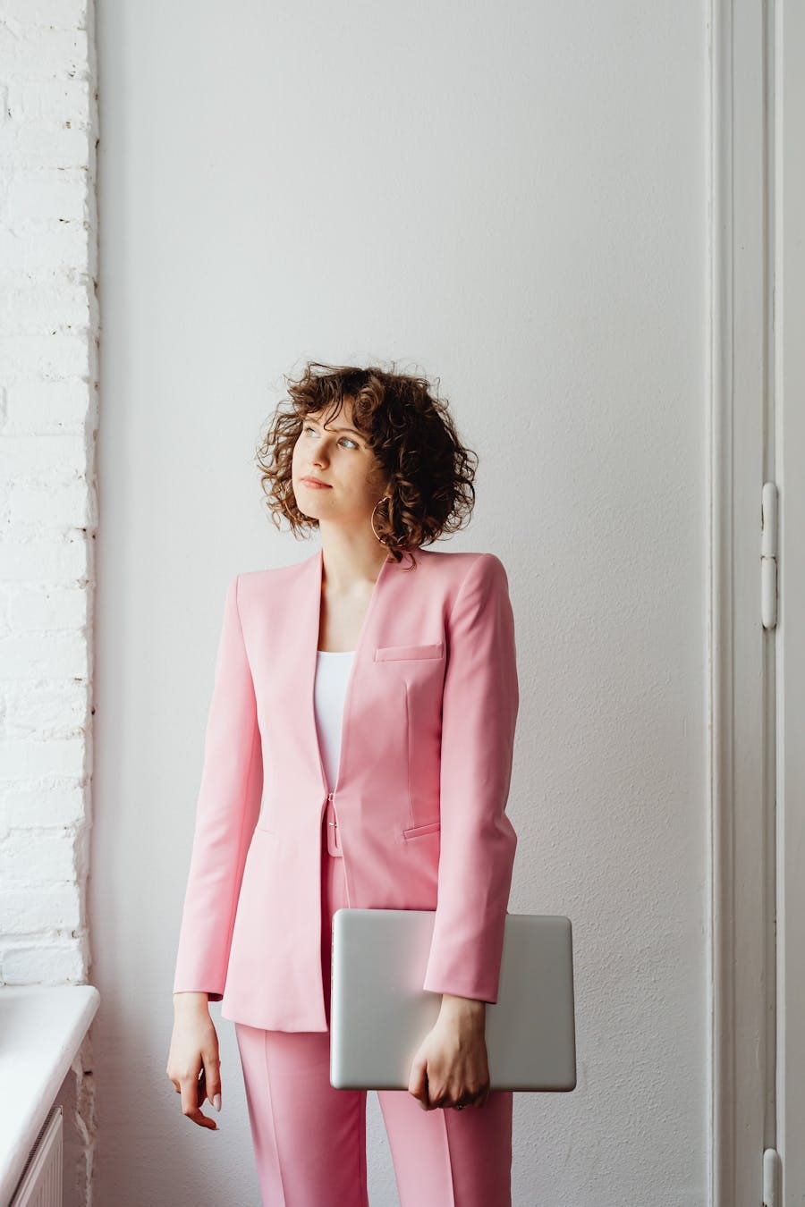 A stylish woman in a pink suit holding a laptop stands indoors with a thoughtful expression.