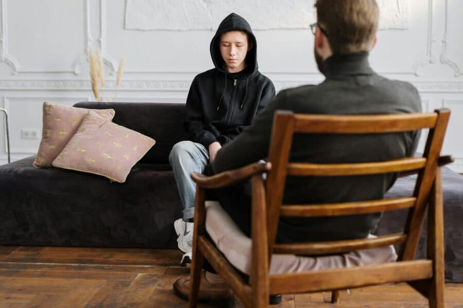 A teenager in hooded sweatshirt talks with a therapist in a modern, minimalistic studio.