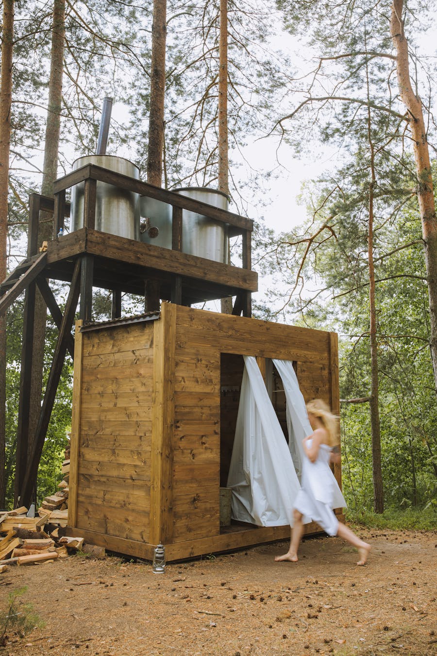 A woman in a white dress approaches an outdoor wooden shower structure in a forest setting.