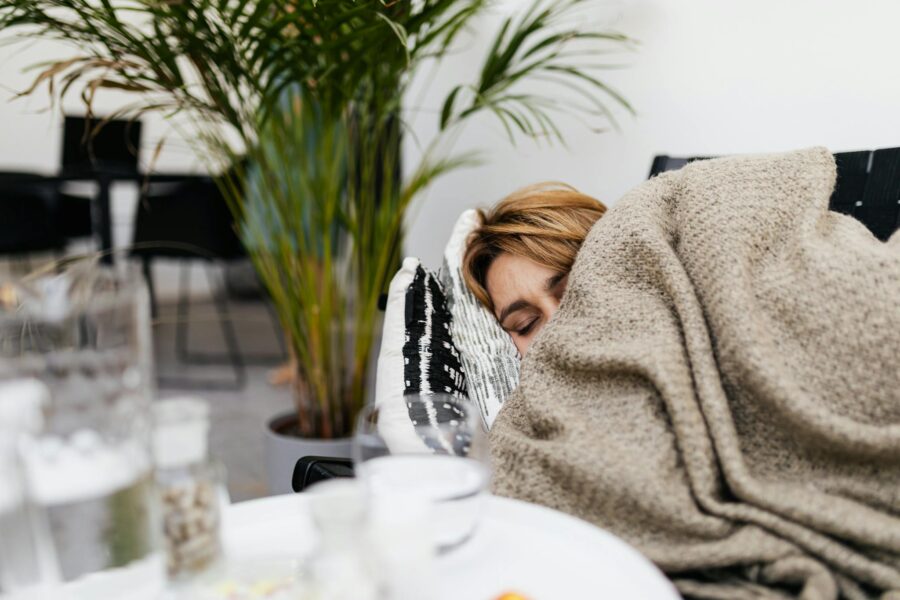 A woman resting on a couch covered by a blanket, indoors with plants.