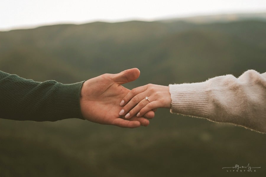 couple holding hands across mountain background with woman wearing engagement ring