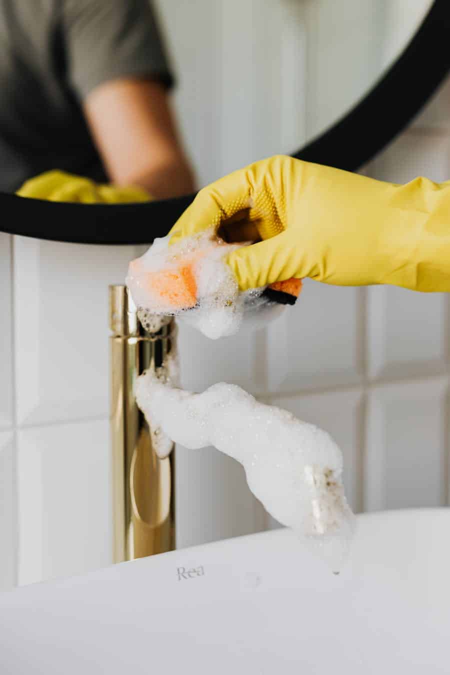 Close-up of a hand in a yellow glove cleaning a bathroom faucet with foam.