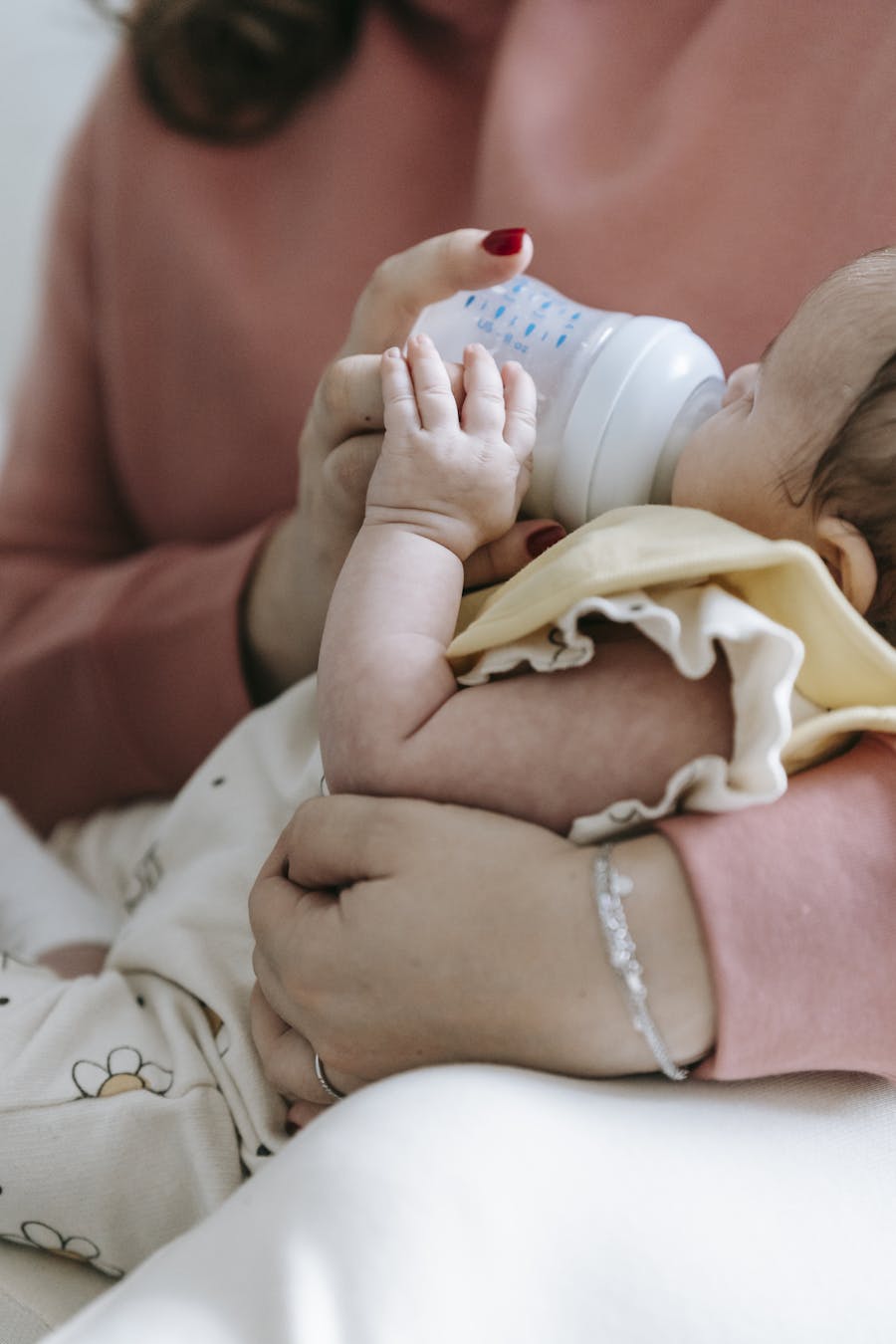 Crop unrecognizable mother in casual wear feeding crop adorable infant with bottle in arms while sitting in light room at home