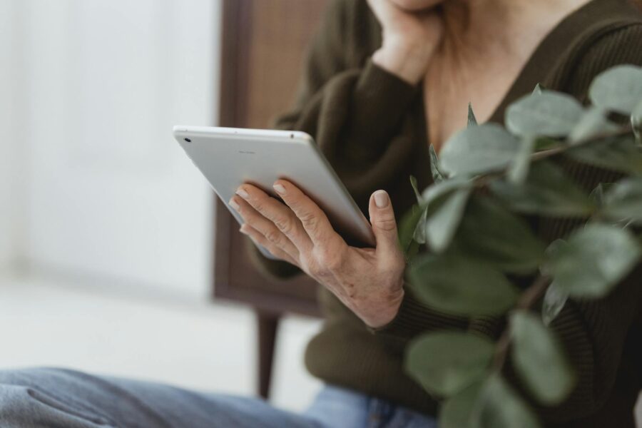 Crop unrecognizable senior female touching face while sitting with cellphone at home against plant on blurred background