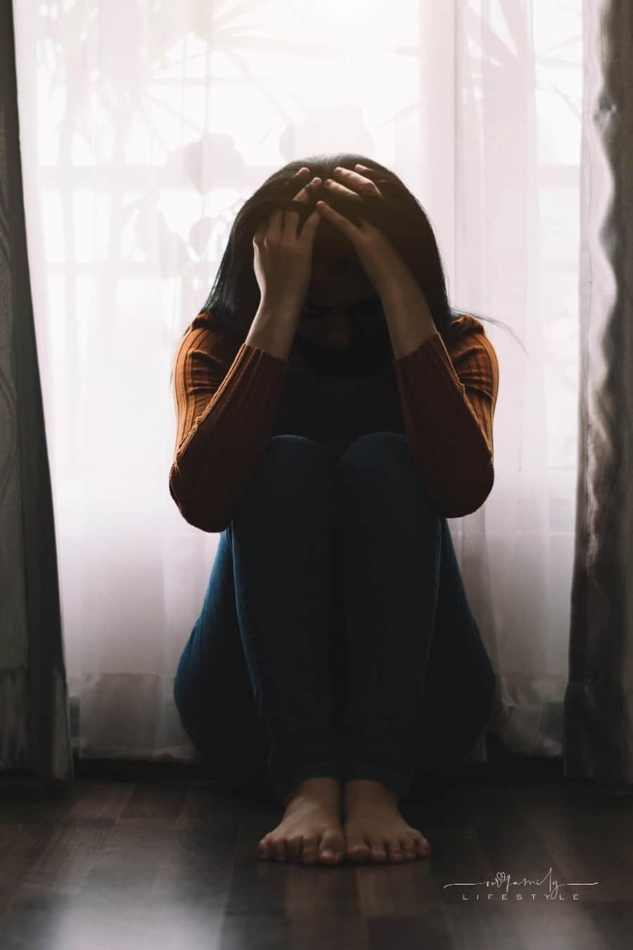 young female with head in hands, depressed in front of sheer white curtain