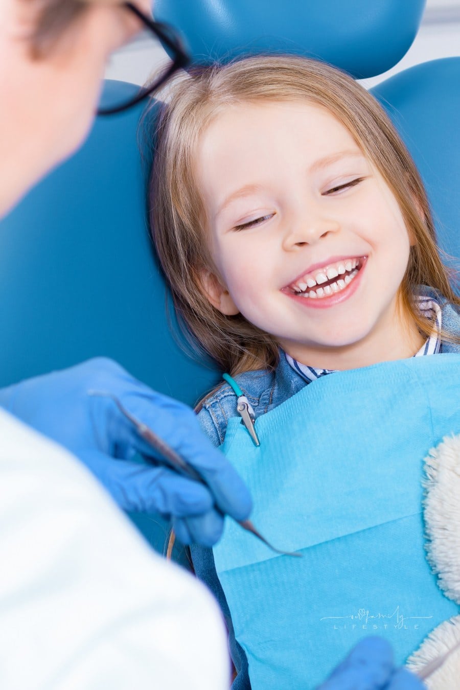 Little cute smiling girl is sitting in dental chair in clinic, office. Doctor is preparing for examination of child teeth with tools, amusing patient with bear toy. Visiting dentist with children.
