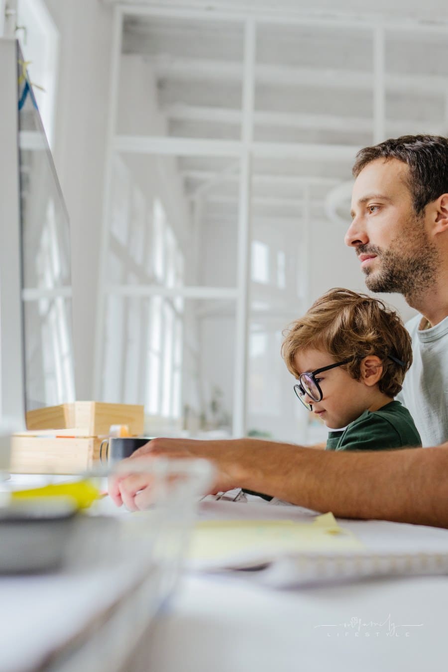 Photo of a cute little boy wearing glasses and his father spending time together in his home-based office; the daily routine of a modern father.