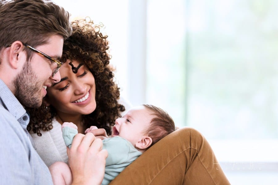 Happy young woman and her husband smile at their sweet newborn baby boy. They are relaxing in their home.