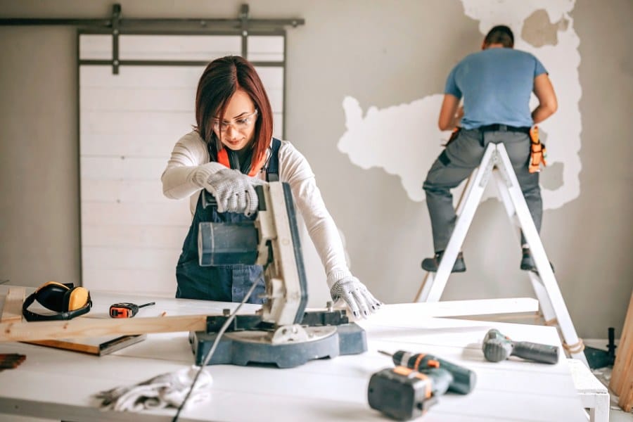 woman cutting boards with a saw while a man works on wall of house behind her