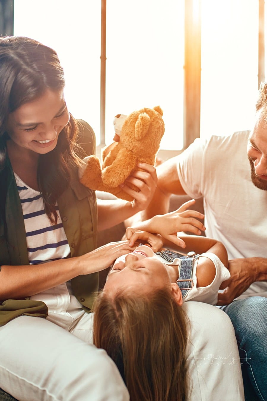 Happy dad and mom with their cute daughter and teddy bear hug, play and have fun sitting on the sofa in the living room at home.