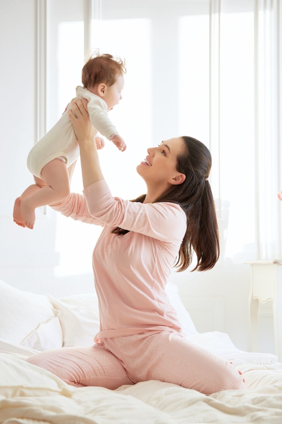 first time mom holding baby up, smiling at her, in bed
