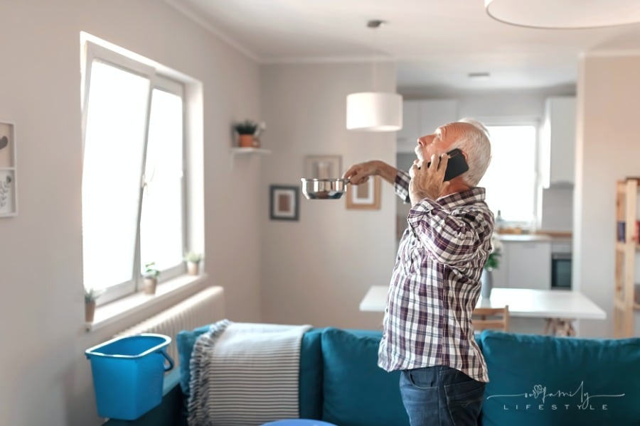 elderly man talking on phone looking up at ceiling holding silver pot to catch water leaking from roof