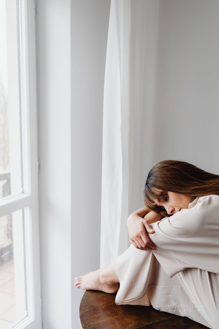 Sad Woman sitting on Table near Window with head on knees