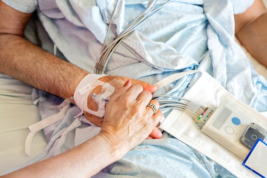 wife holding husband's hand as he lays in a hospital bed