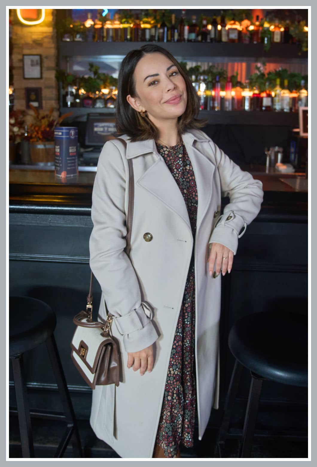 Janel Parrish in character as Jana from the Hallmark movie Haunted Wedding, dressed in a floral dress and cream coat, smiling at a cozy bar setting