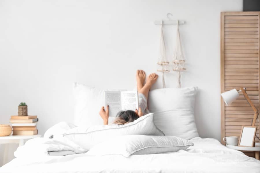 woman lying in bed with feet on wall reading a book