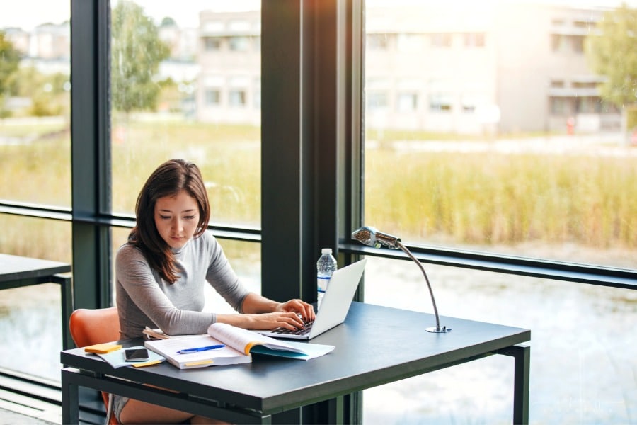 college student taking notes from books in library as she studies