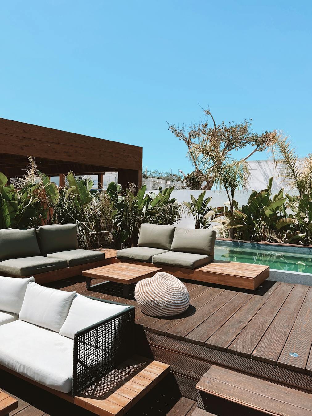 Luxurious outdoor patio with pool, modern furniture, and lush plants under a clear sky.