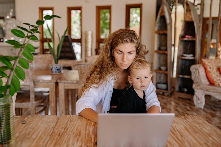 Mother and child sit at a wooden table with a laptop indoors, focusing together.