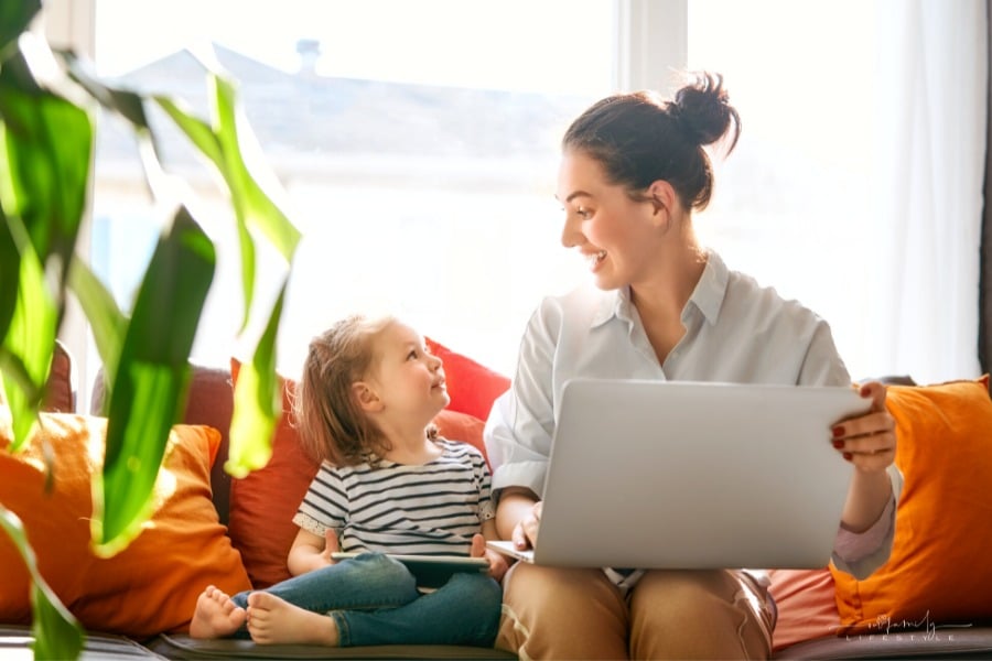 mom sitting on couch with laptop in hand and smiling at young daughter beside her