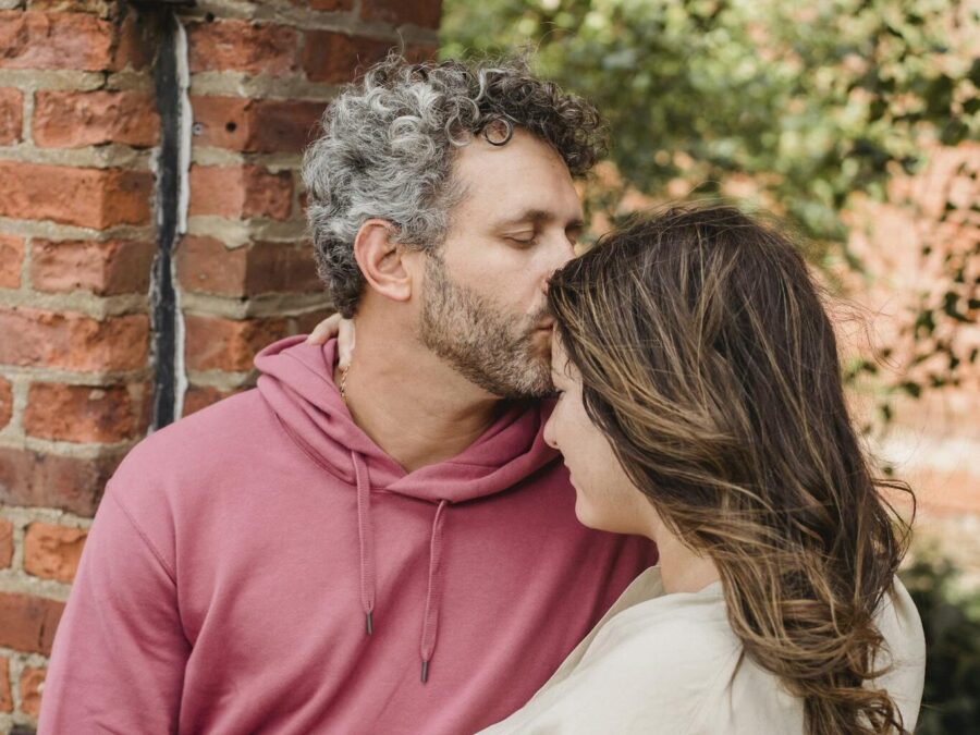 Romantic man in stylish clothes touching forehead of pregnant wife with closed eyes while resting together in green park near