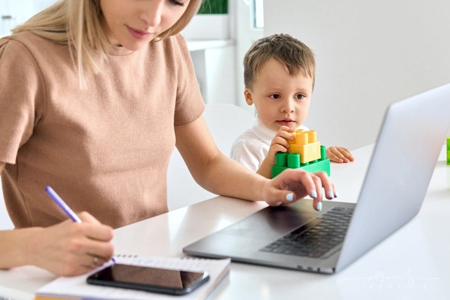 mom writing on notepad while working at laptop with young son nearby
