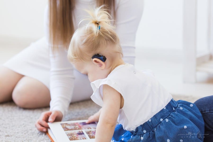 toddler girl wearing a hearing aid while looking at book with mom