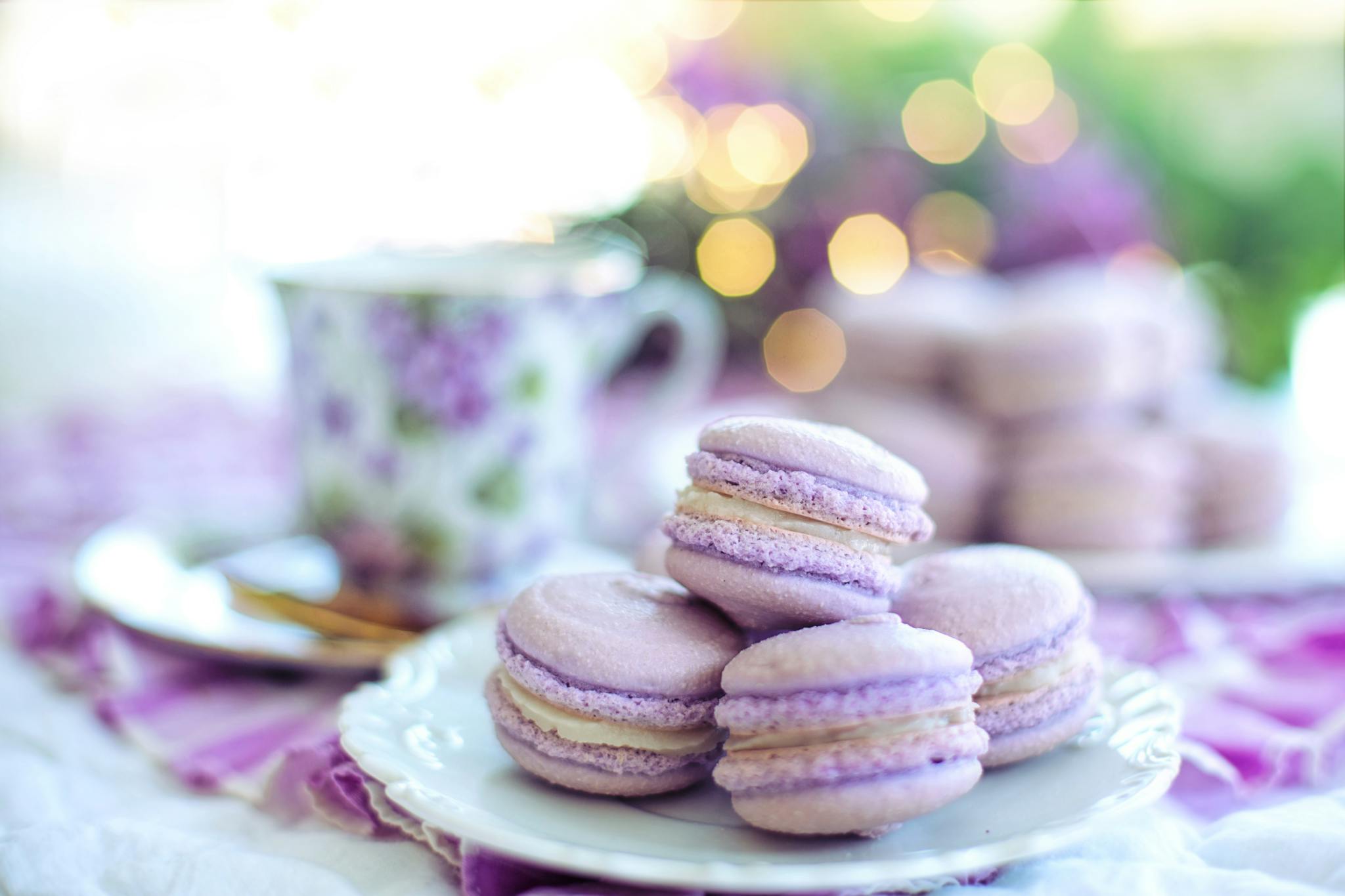 Soft purple macarons on a white plate with a floral teacup, surrounded by a warm bokeh background.
