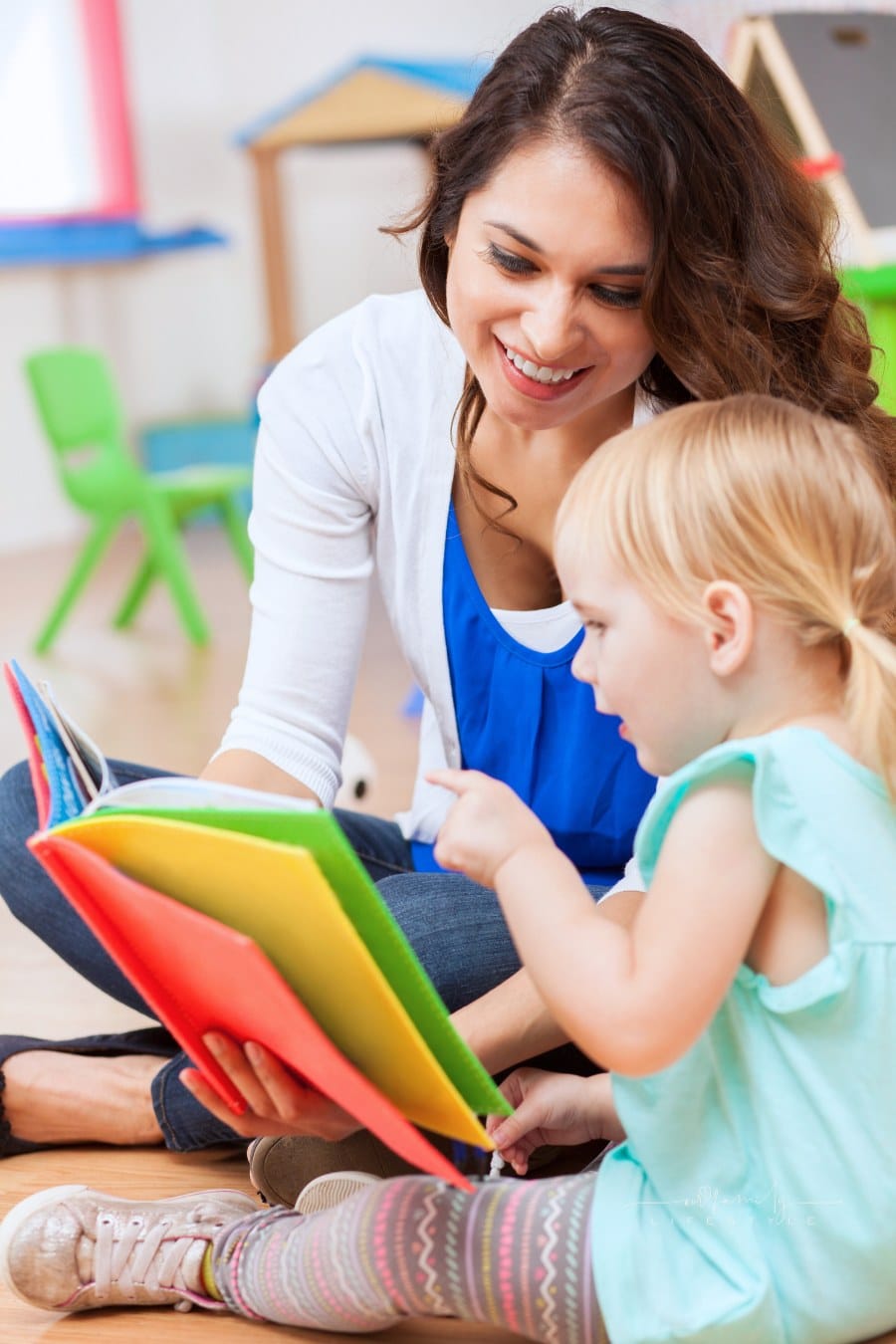 daycare teacher sitting on the floor and reading a colorful book to a preschool girl. The little girl is pointing and interested in the book as the teacher reads to her.
