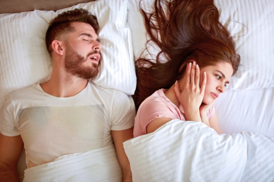 woman covering her ears while man snores in bed behind her