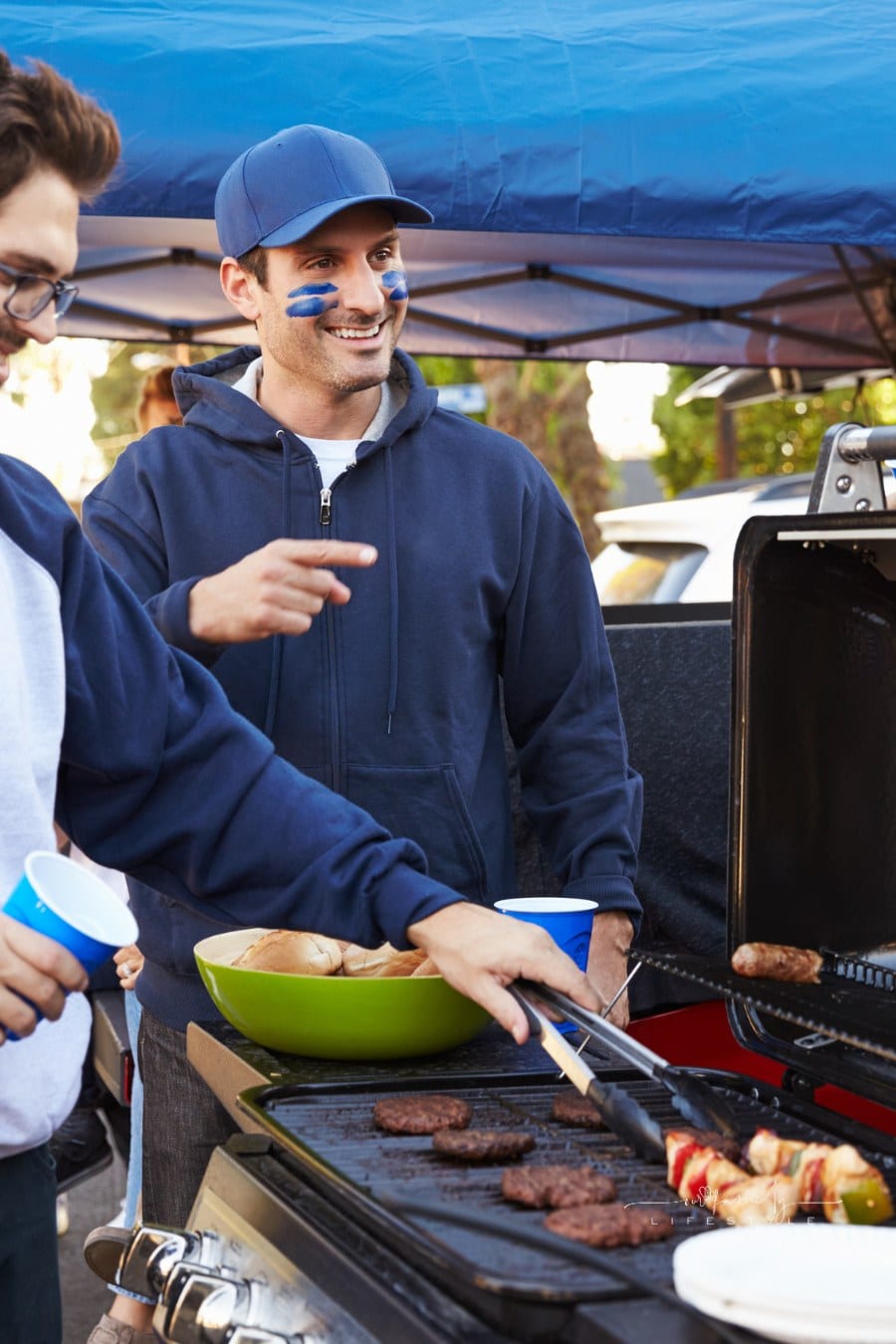 Group of Male Sports Fans Tailgating in stadium parking lot; grilling burgers on back of truck
