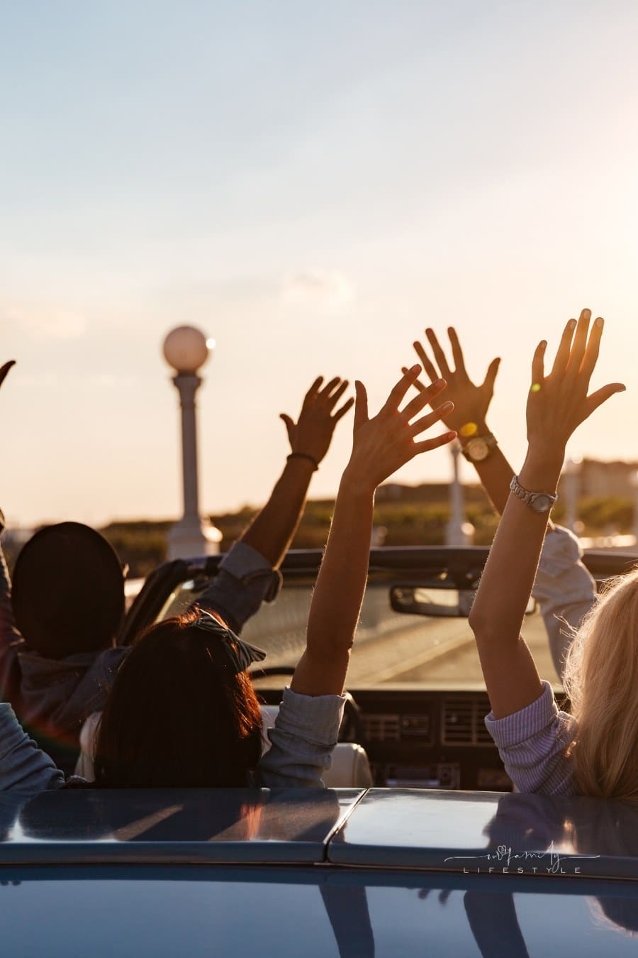 Back view of happy young friends driving cabriolet with raised hands on sunset