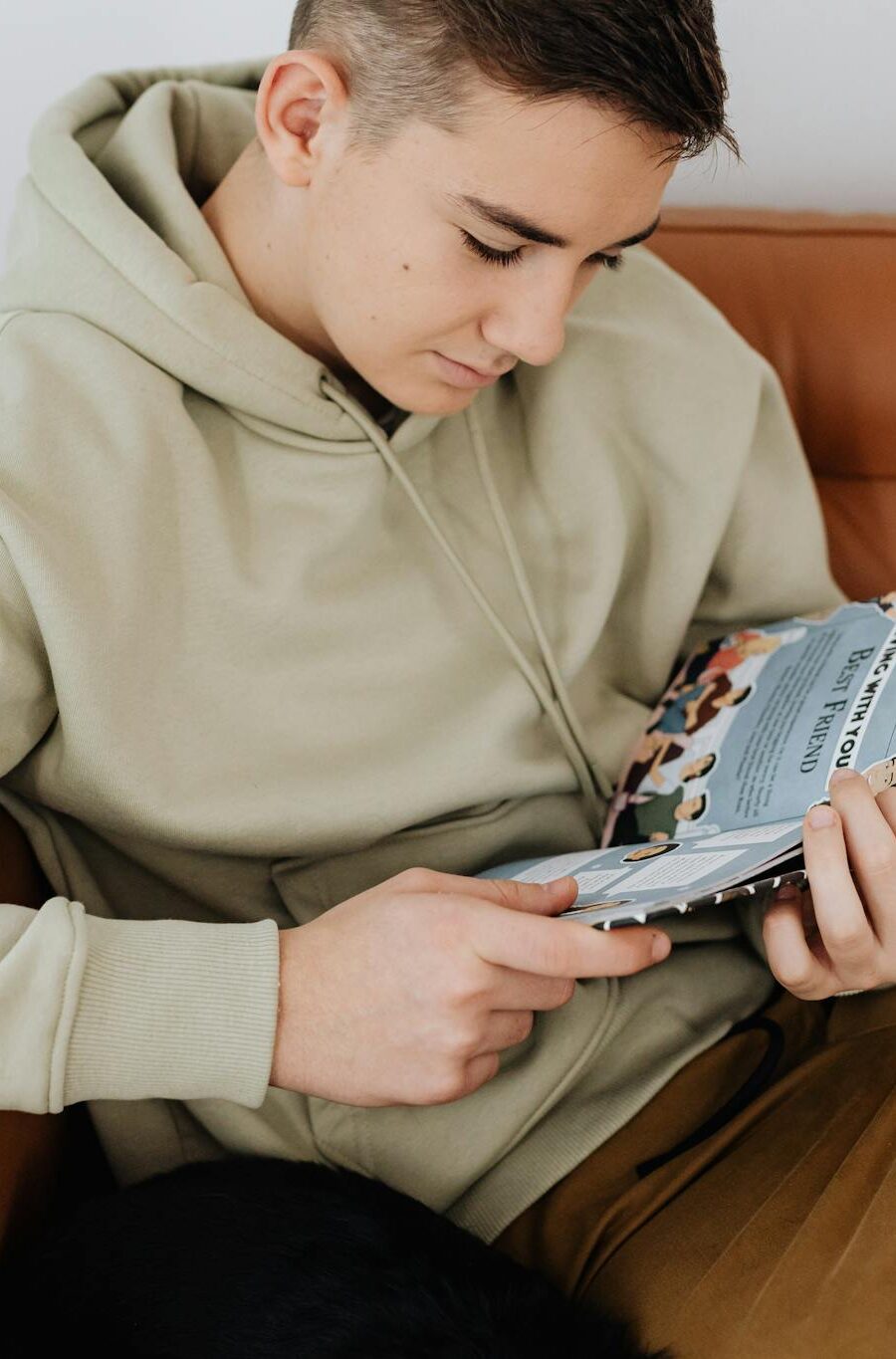 Teenage boy in hoodie engrossed in reading a magazine while sitting comfortably on a couch.