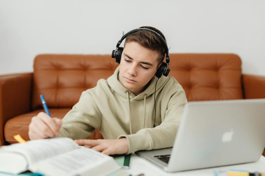 Teenage boy with headphones, focused on studying, using a laptop indoors.