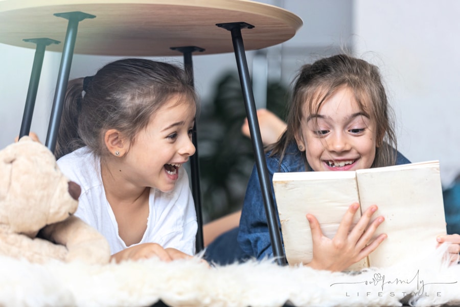 Two little sisters are reading a book lying on the floor on a cozy blanket.