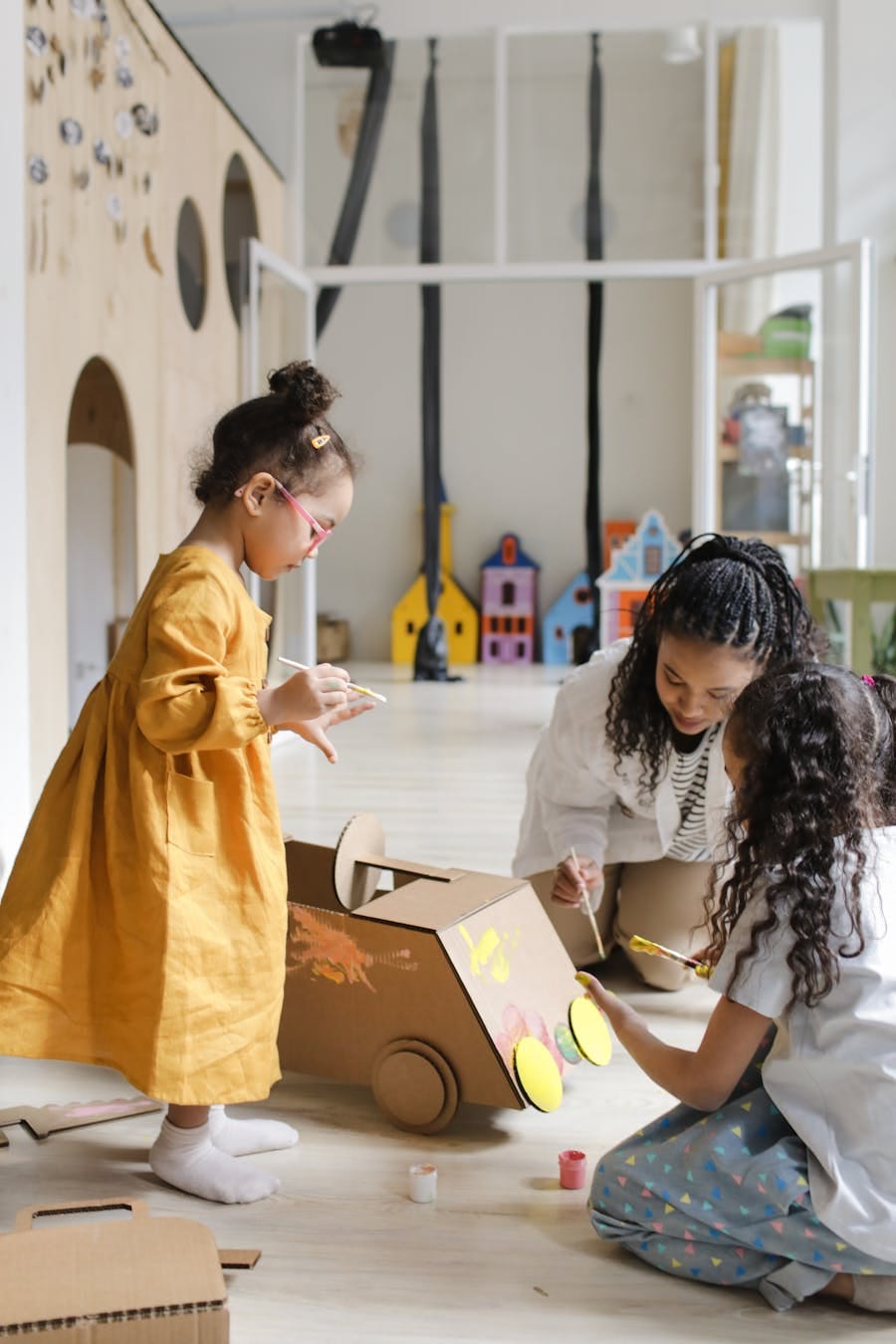 Two young girls painting a cardboard box car indoors, guided by a caregiver in a nurturing environment.