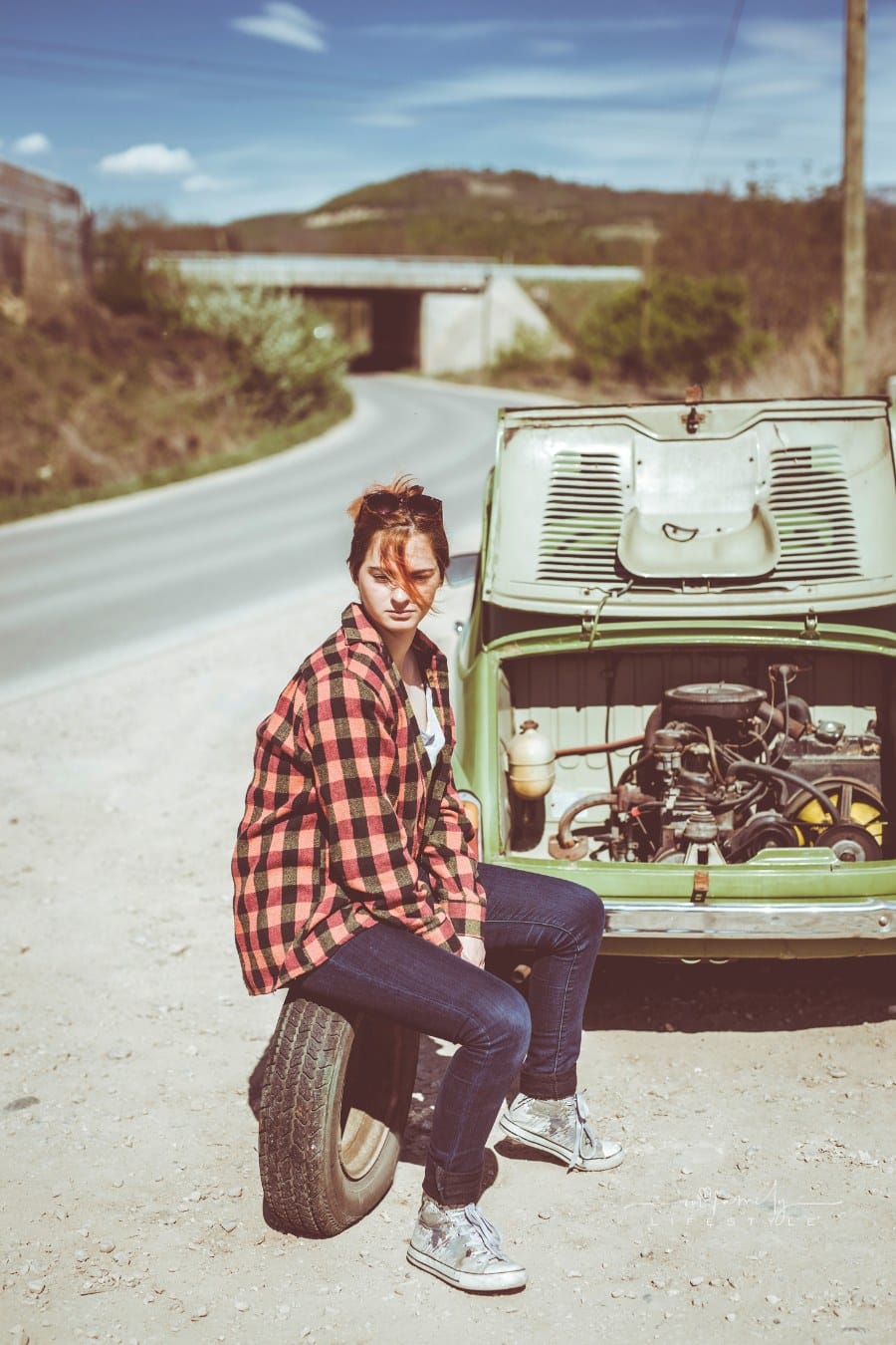 A girl with a spare tire next to her broken down green mini car