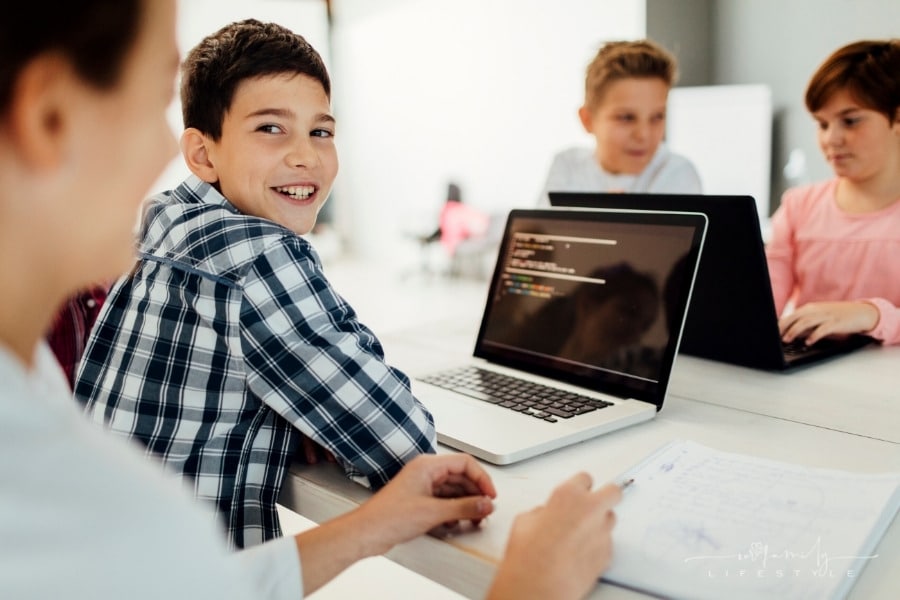 young boy smiling at camera with laptop open while learning to code