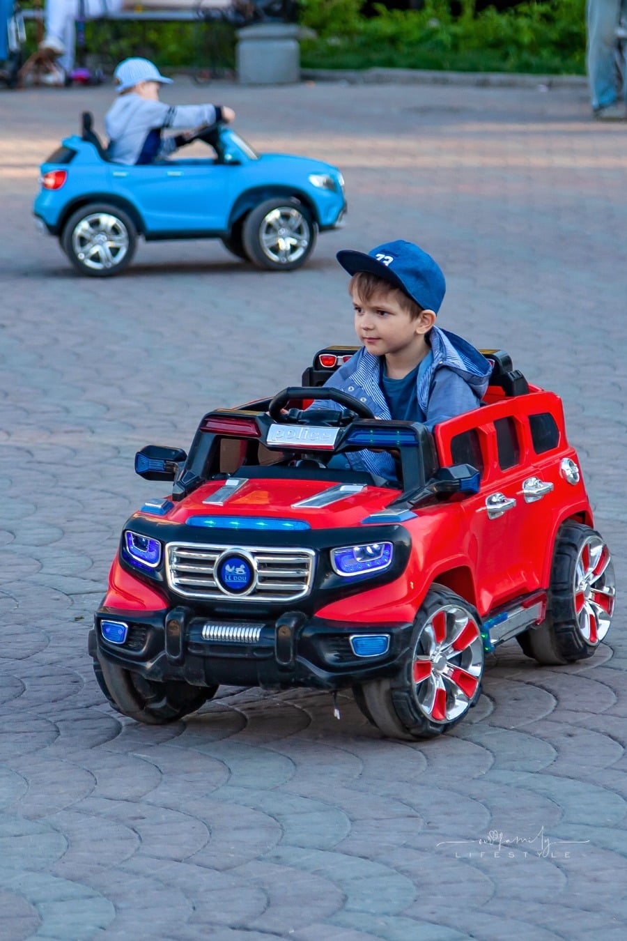 Two little boys ride on red and blue electric cars while walking with their parents in a city park with green trees, laughing and smiling
