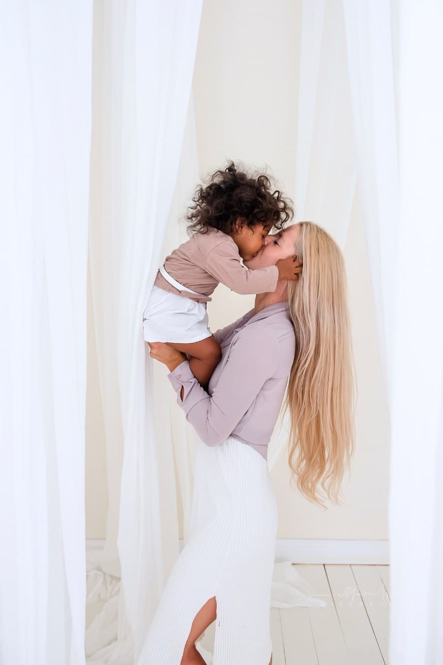 Young blond Caucasian mother holding African American curly daughter in arms among the white cuts of floating light tulles. Adoption, mixed ethnic family