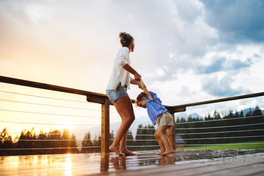 Mother with Small Daughter Playing in Rain on Patio of Wooden Cabin