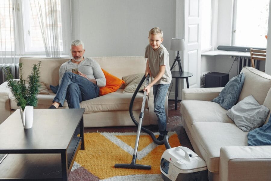 A boy vacuums the living room while an older man sits on the sofa using a phone.
