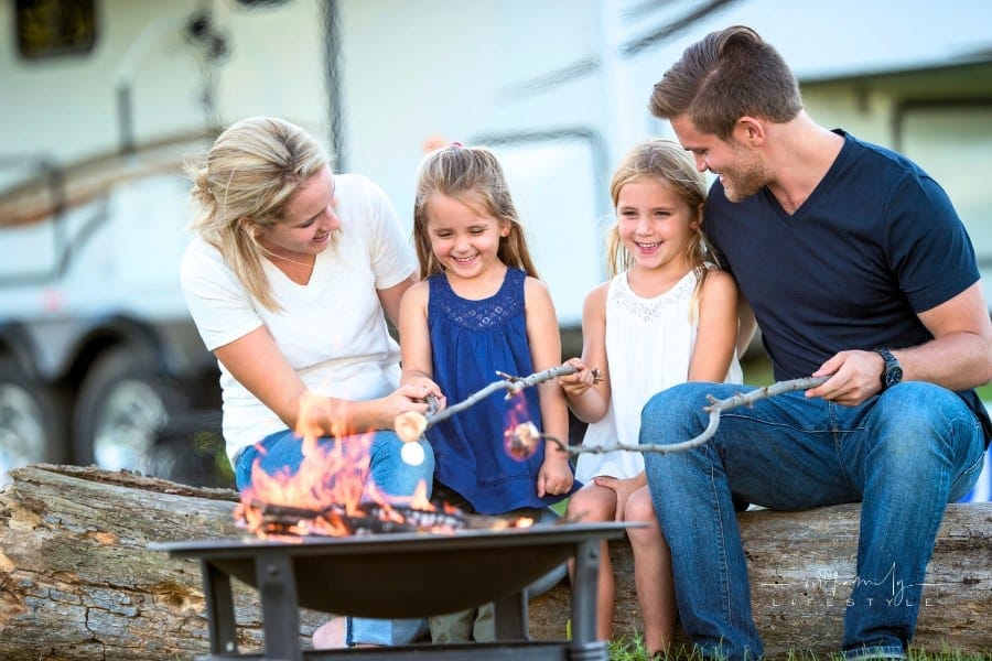 A family of four are roasting marshmallows together around a campfire while on vacation. Their RV is parked in background.